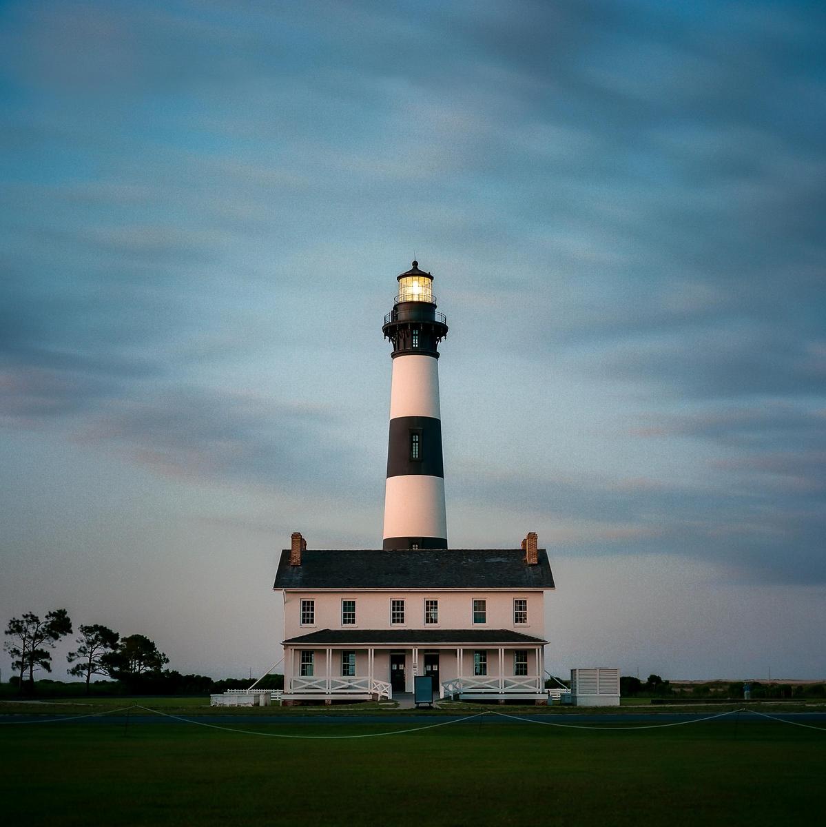 A black and white lighthouse with an illuminated top stands next to a white building under a cloudy sky at dusk.