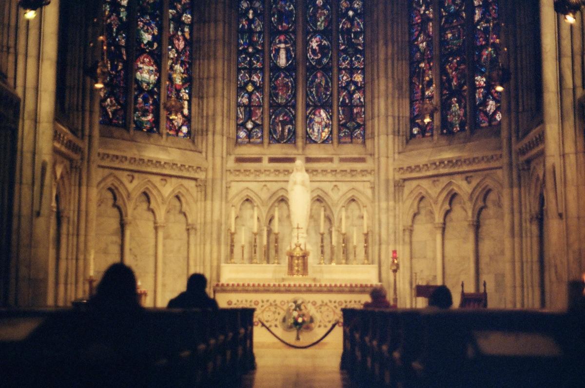 People sit in pews facing an altar with a statue and stained glass windows in a church.