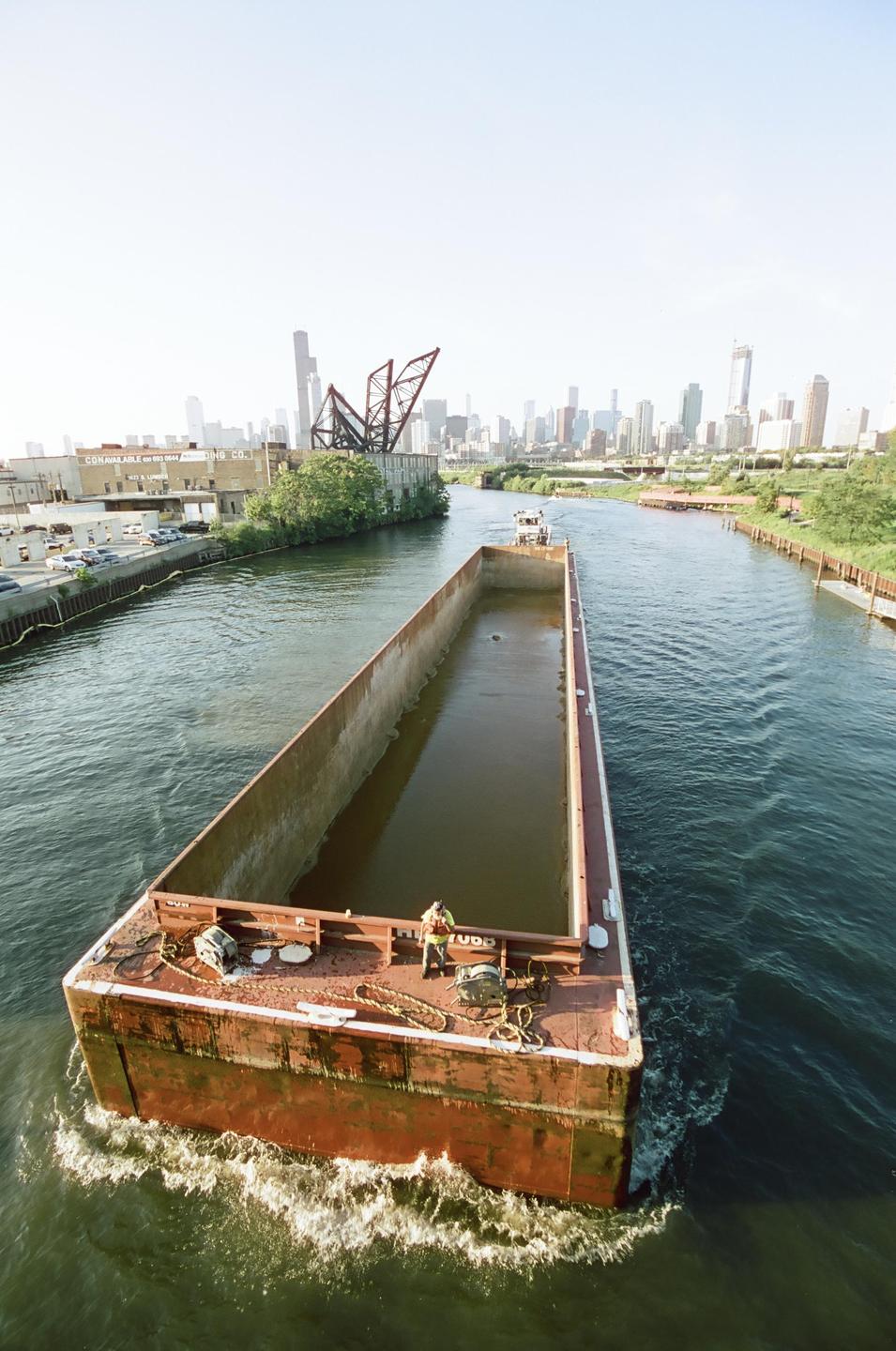 A large barge with a man on board travels down a river with a city skyline in the background.