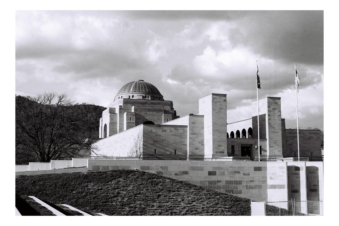 A large domed building with flags flying in front of it stands on a hill with trees and a cloudy sky in the background.