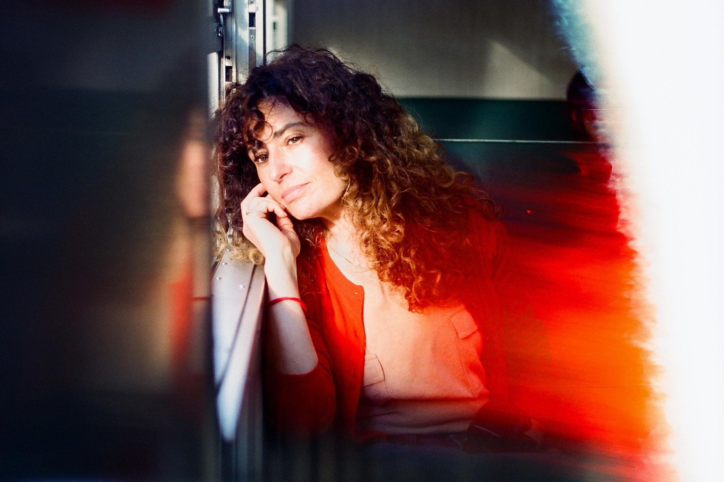 A woman with curly hair rests her head on her hand while sitting indoors, with a bright red light leak on the right side of the image.