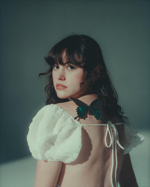A woman with dark hair looks over her shoulder, with a green butterfly on her back.
