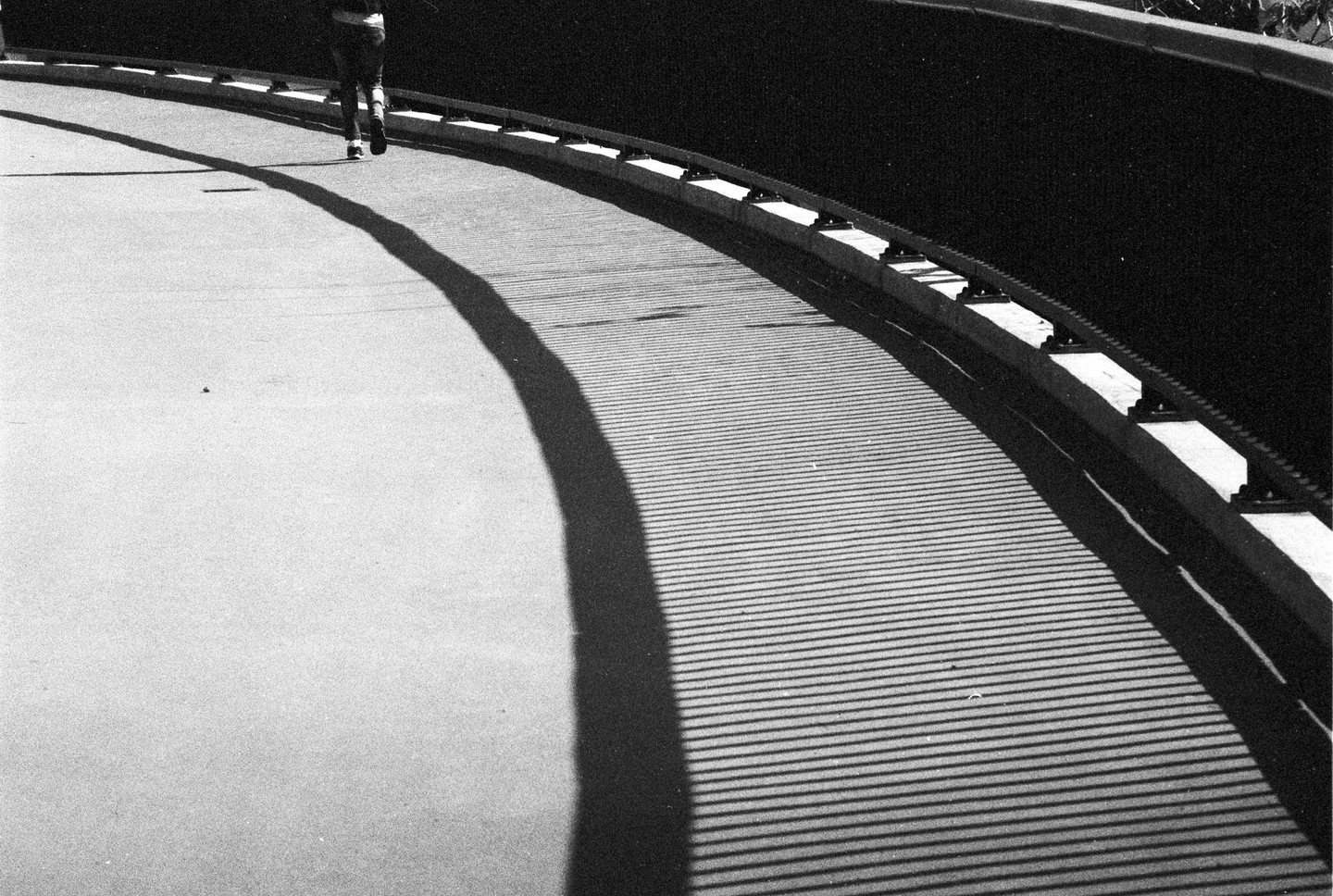 A person walks on a curved path with a long shadow stretching across the ground, which is patterned with parallel lines of shadow.