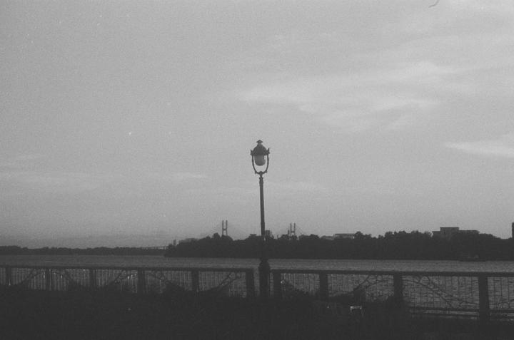 A street lamp stands in front of a body of water with a distant bridge and trees on the horizon.