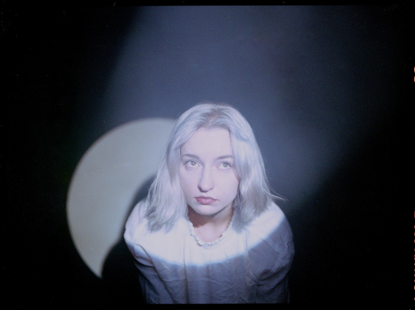A woman with light hair looks up, illuminated by a spotlight against a dark background.