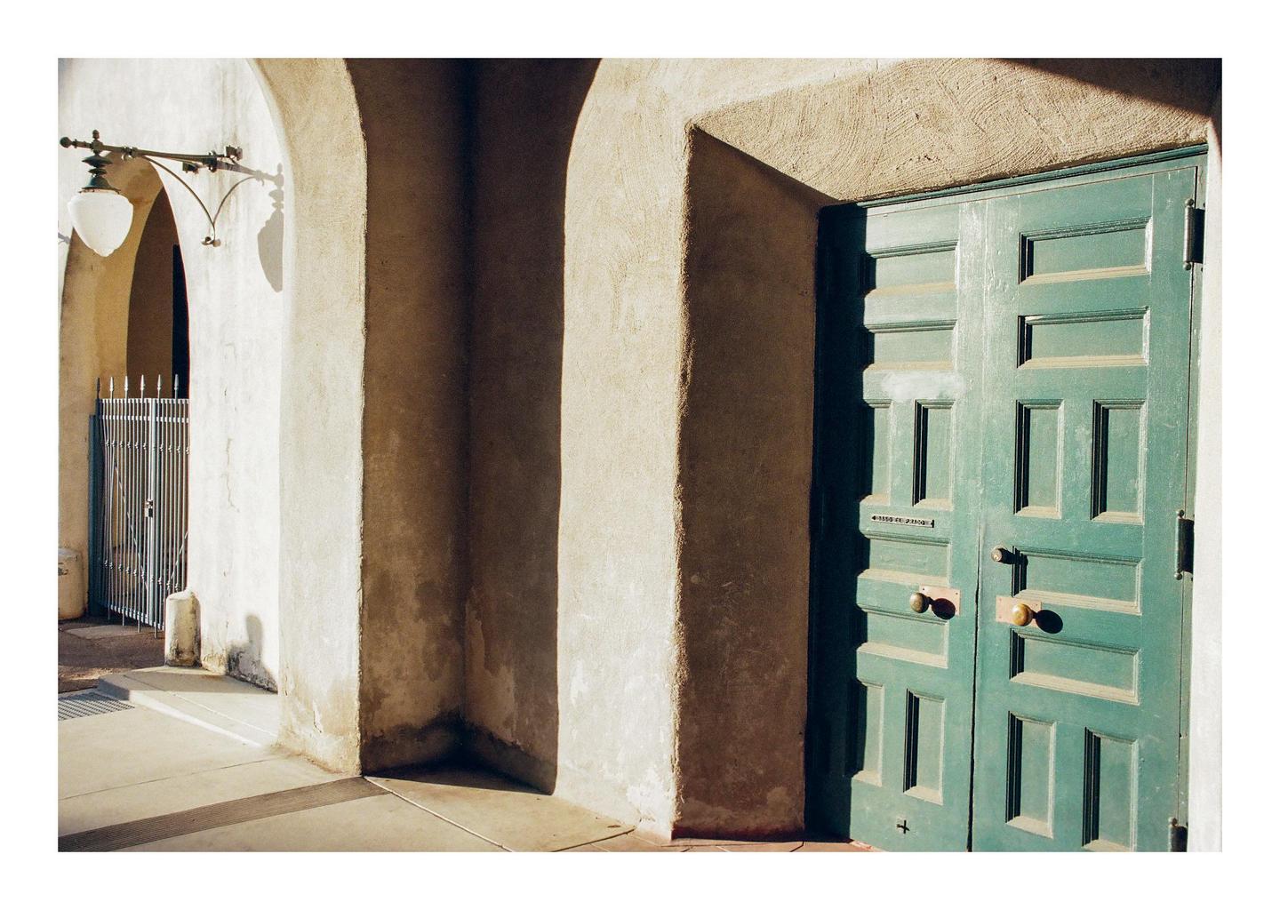 A green door is next to a series of arches with a street lamp and a gate in the background.