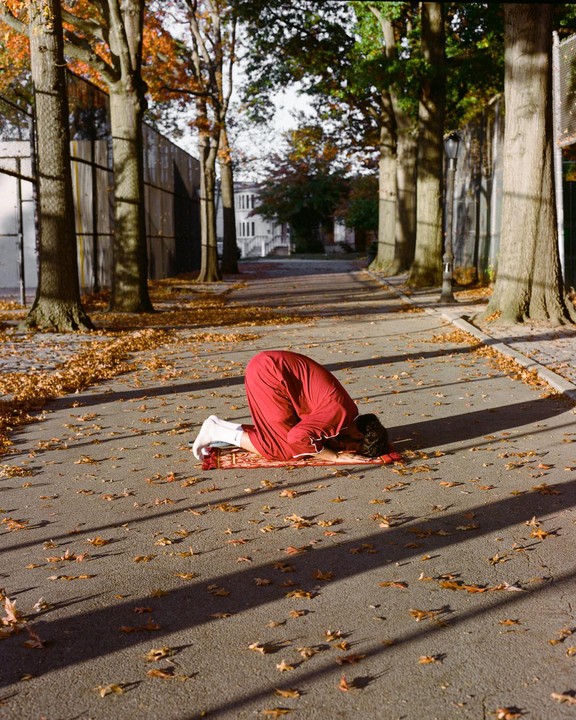 A person in red clothing kneels on a prayer mat on an asphalt path covered with autumn leaves, with trees and buildings in the background.