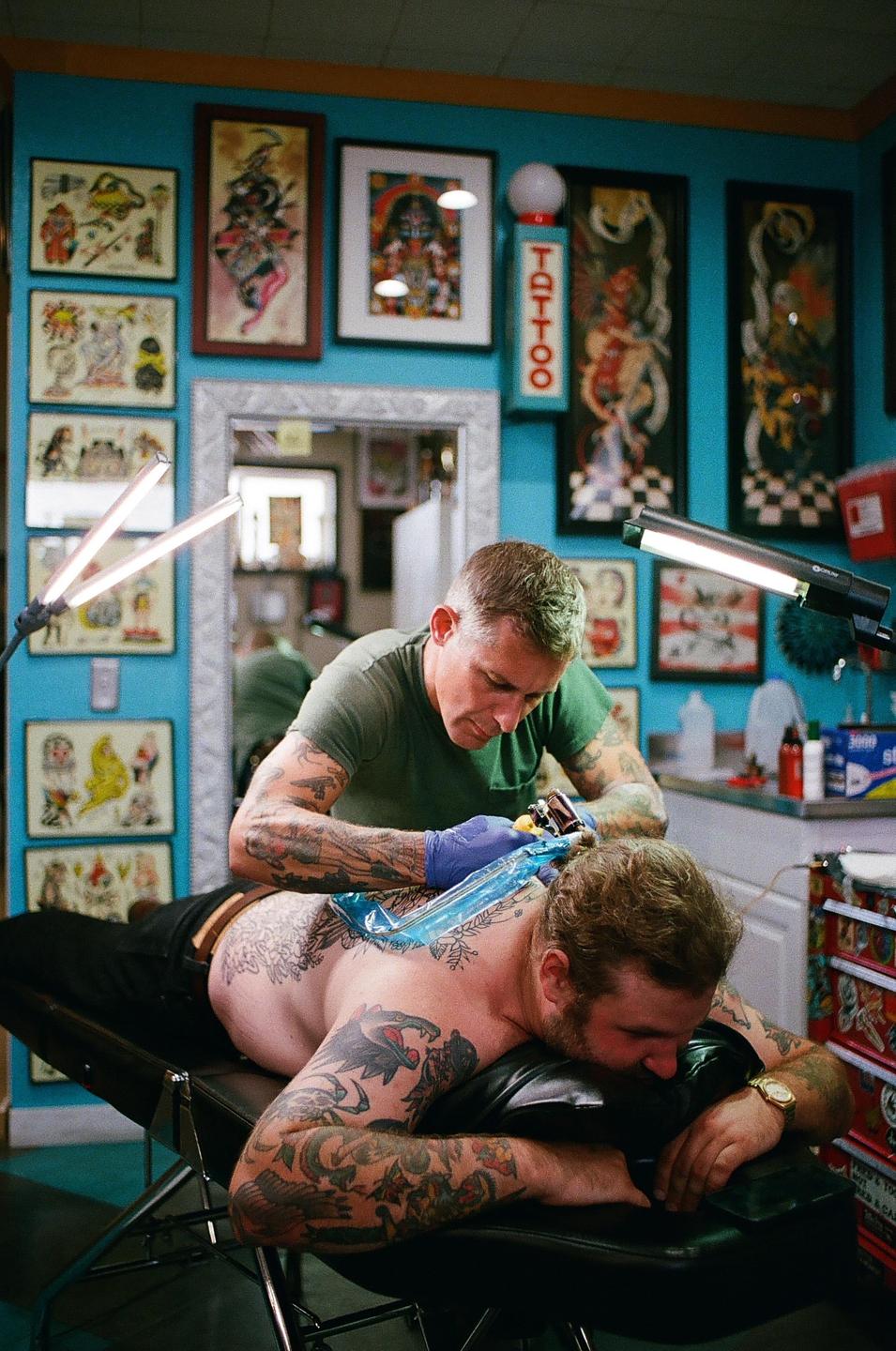 A man with a beard lies on his stomach while another man tattoos his back in a room with blue walls covered in framed tattoo designs.