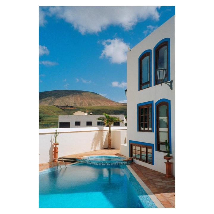 A white building with blue trim stands next to a swimming pool, with a mountain in the background under a blue sky.