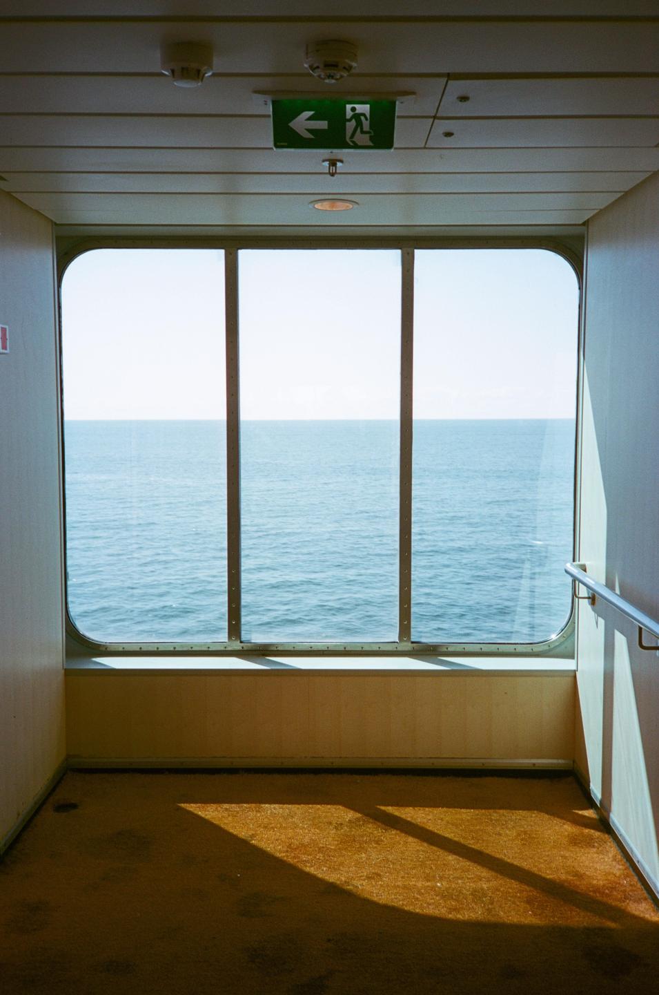 A window on a cruise ship looks out onto the open ocean with sunlight casting shadows on the carpeted floor.