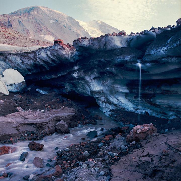 A stream flows from a dark ice cave with a small waterfall, surrounded by rocky terrain and mountains in the background.
