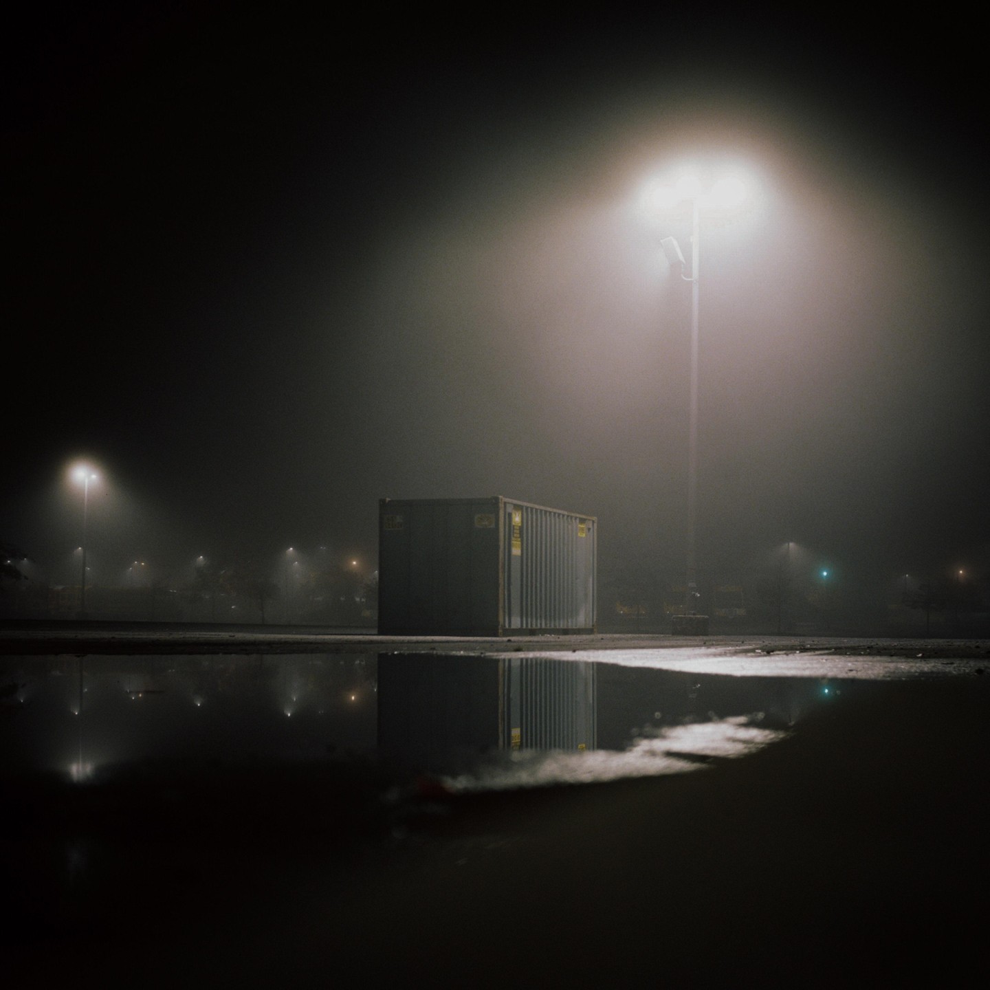 A shipping container sits in a parking lot at night, illuminated by a street light and reflected in a puddle.