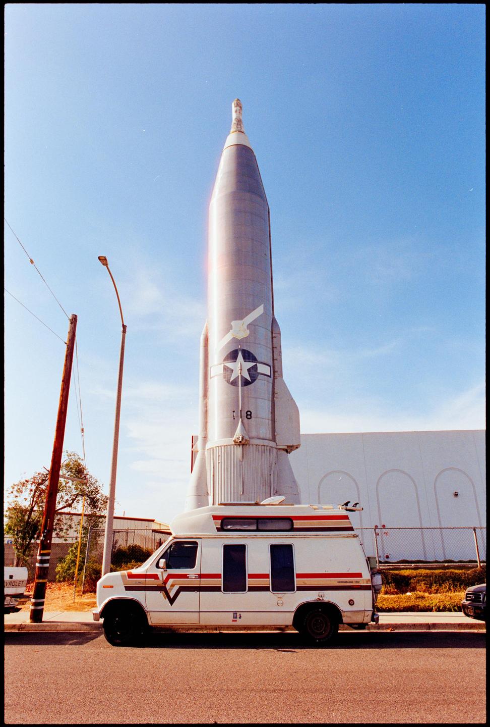 A white van with surfboards on its roof is parked in front of a large rocket under a clear sky.