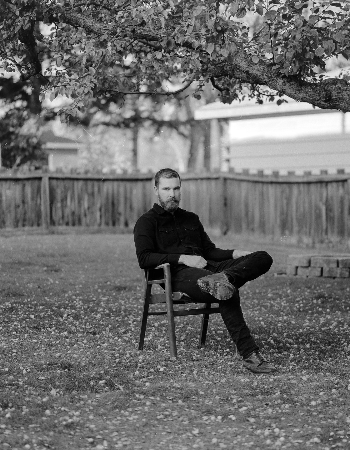 A man with a beard sits on a chair in a grassy yard under a tree.