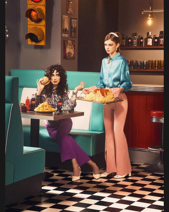 Two women are in a diner, one holding a burger and the other carrying a tray of spaghetti.