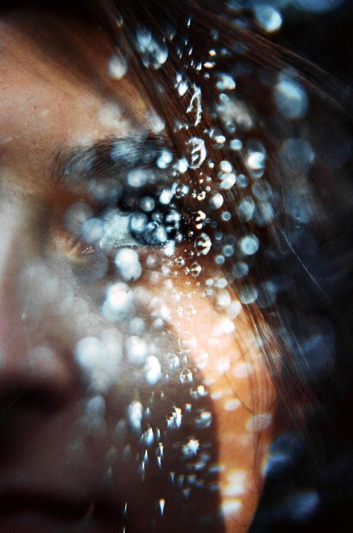 A woman's face is partially obscured by many bright, out-of-focus water droplets.