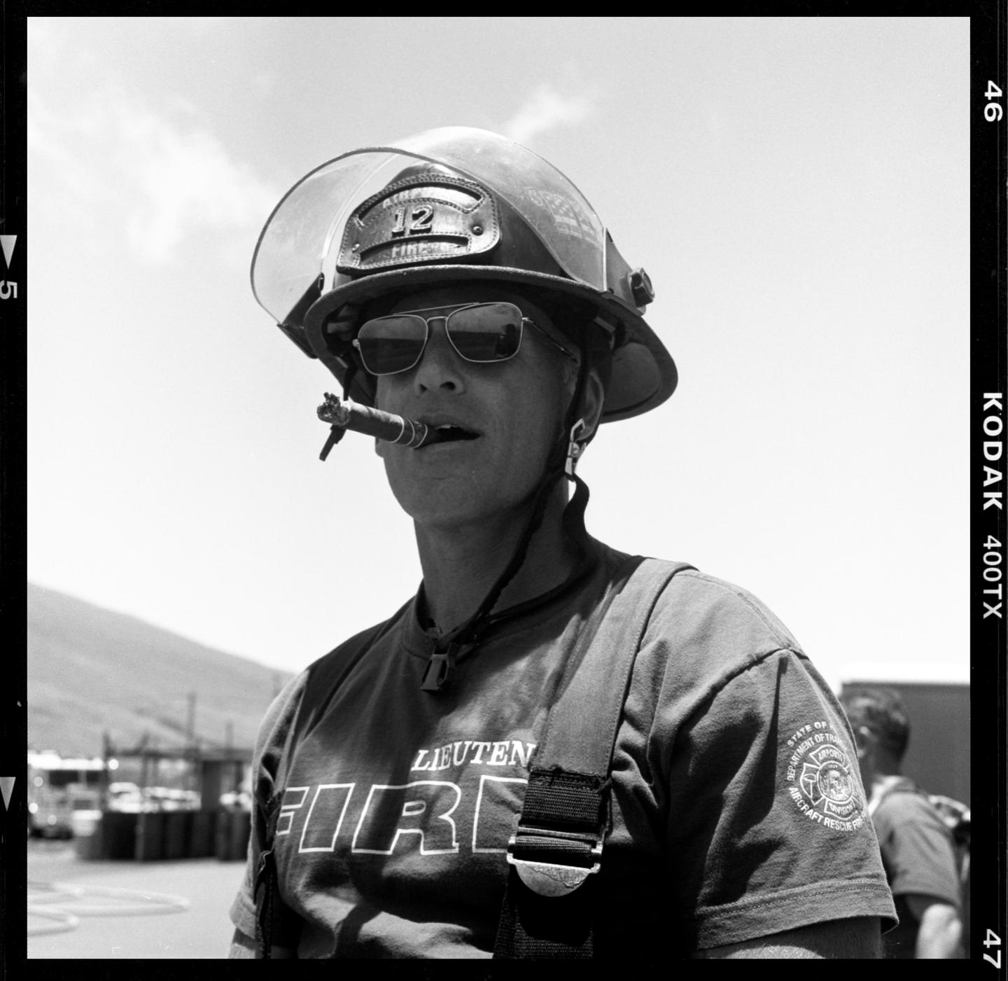 A man wearing a firefighter helmet and sunglasses smokes a cigar.