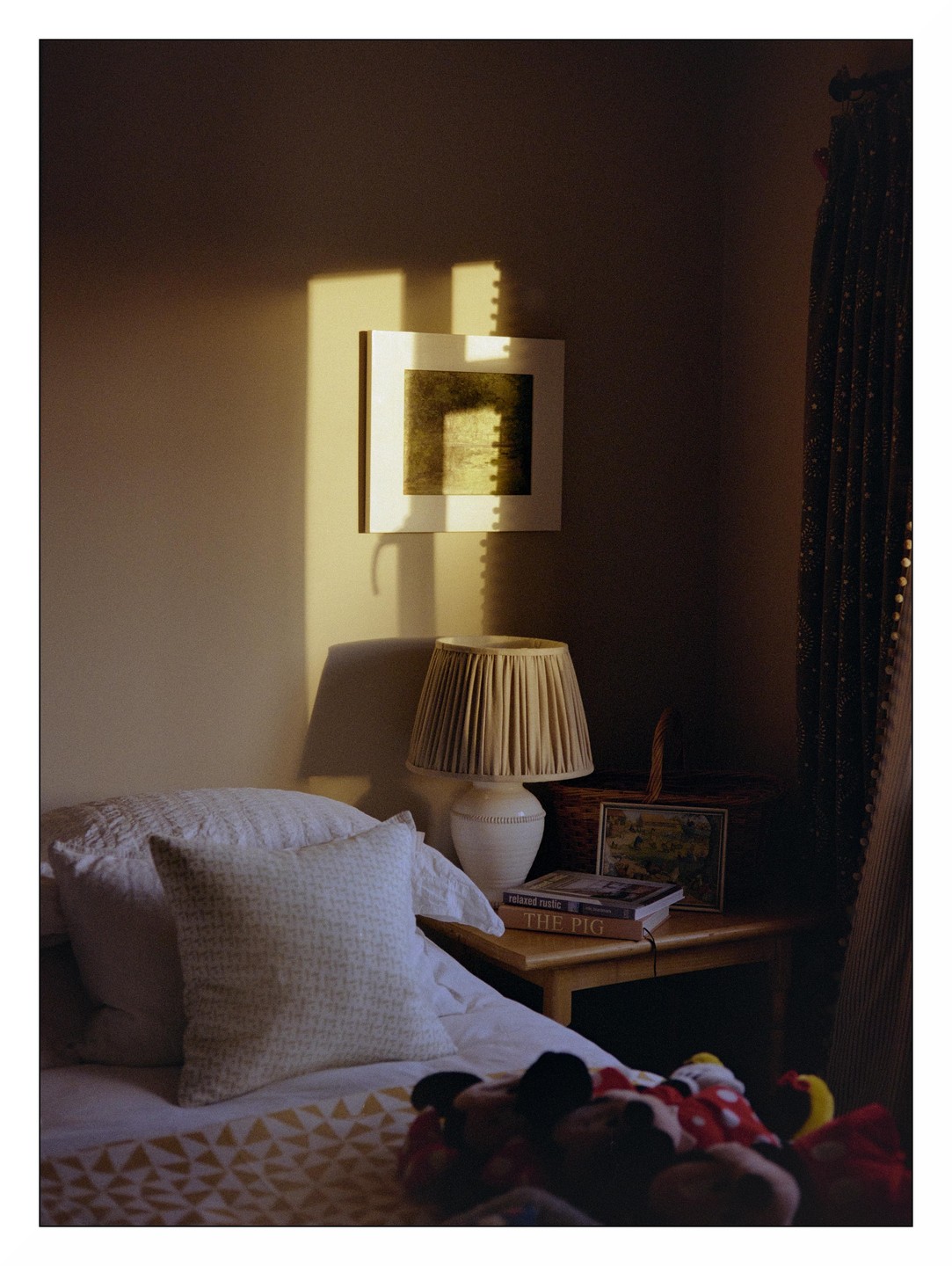 Sunlight streams into a bedroom, illuminating a framed picture on the wall and a bedside table with a lamp and books.
