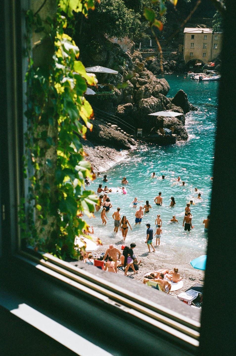A view from a window shows a crowded beach with people swimming and sunbathing in turquoise water next to a rocky cliff with buildings.