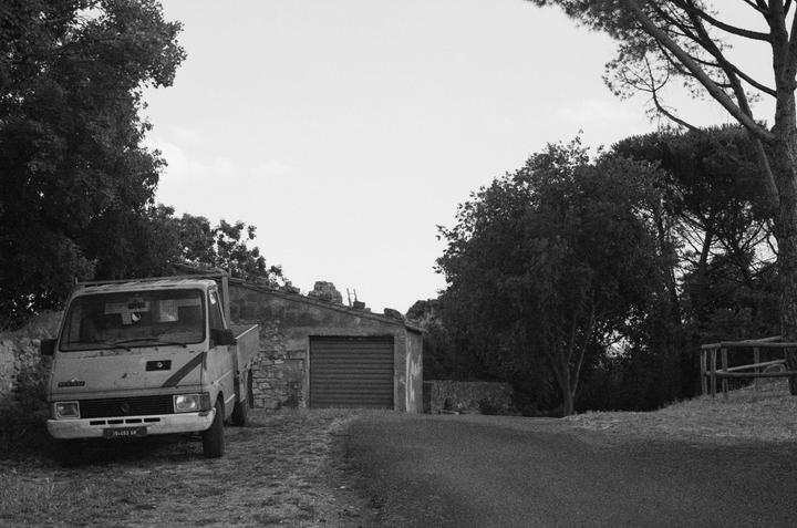 A small truck is parked on a grassy area next to a paved road, with a stone building and trees in the background.