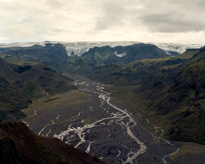 A wide river with many tributaries flows through a valley surrounded by mountains with snow-capped peaks under a cloudy sky.
