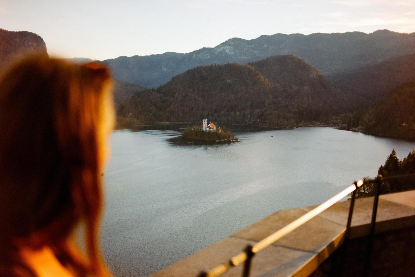 A woman looks out over a lake with a church on an island, surrounded by mountains.