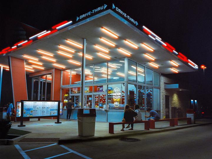 A group of people are gathered outside a brightly lit drive-thru restaurant at night.