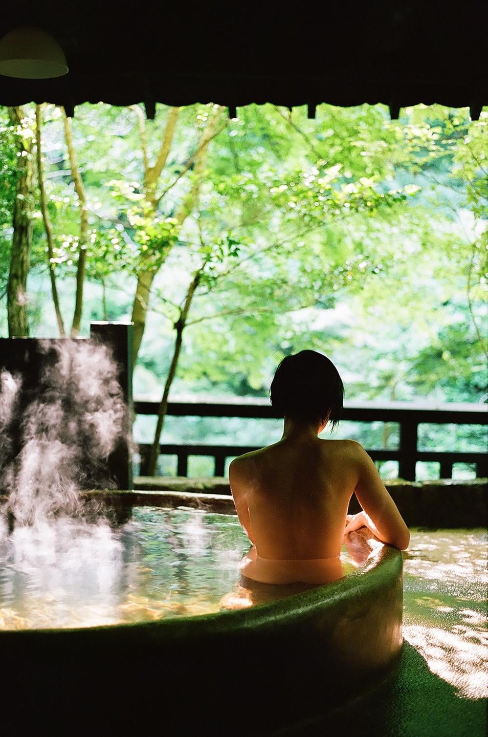 A woman sits in a steaming outdoor bath, looking out at a lush green forest.
