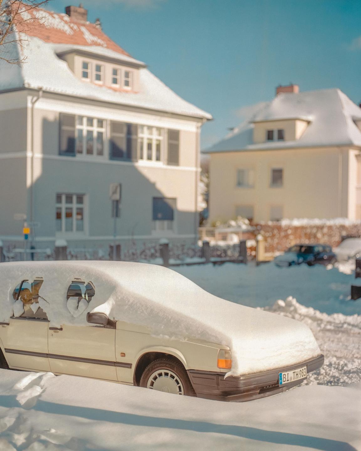 A white car is covered in snow on a street with houses in the background.
