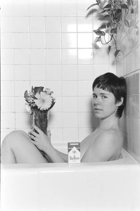 A woman sits in a bathtub holding a vase of flowers, with a pack of cigarettes beside her.
