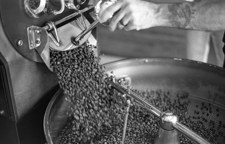 A person's arm is visible as coffee beans pour from a roaster into a cooling tray.