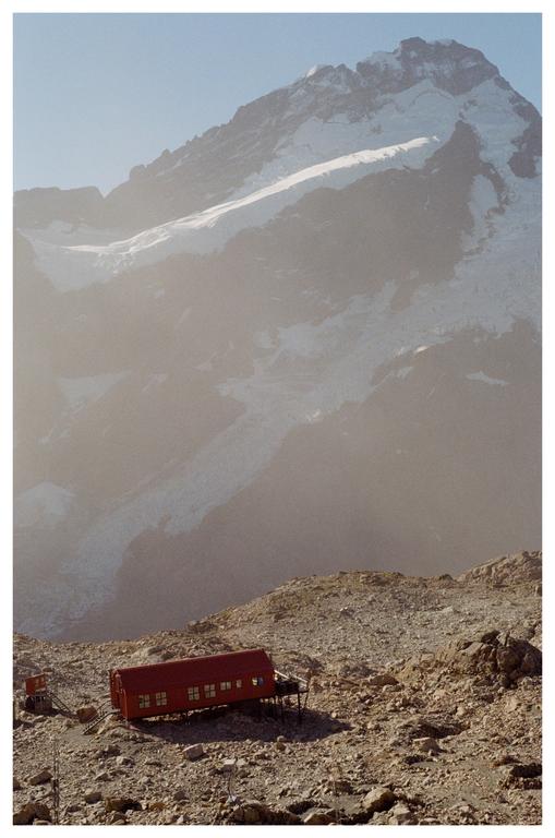 A red hut sits on a rocky mountain slope with a snow-capped mountain in the background.
