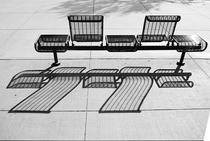 A black and white photo shows a metal bench casting a long shadow on a concrete surface.