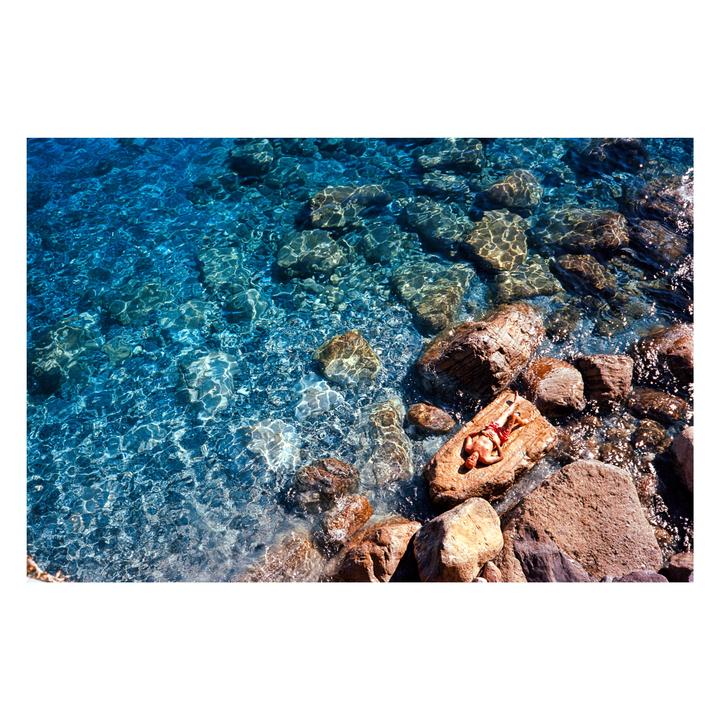 A man sunbathes on a large rock at the edge of clear blue water with many submerged rocks.