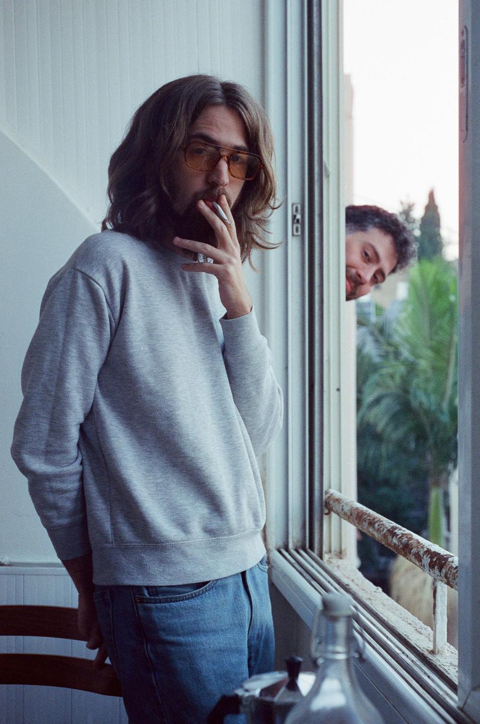 A man with long hair and a beard smokes a cigarette while looking out a window, with another man peeking in from outside.