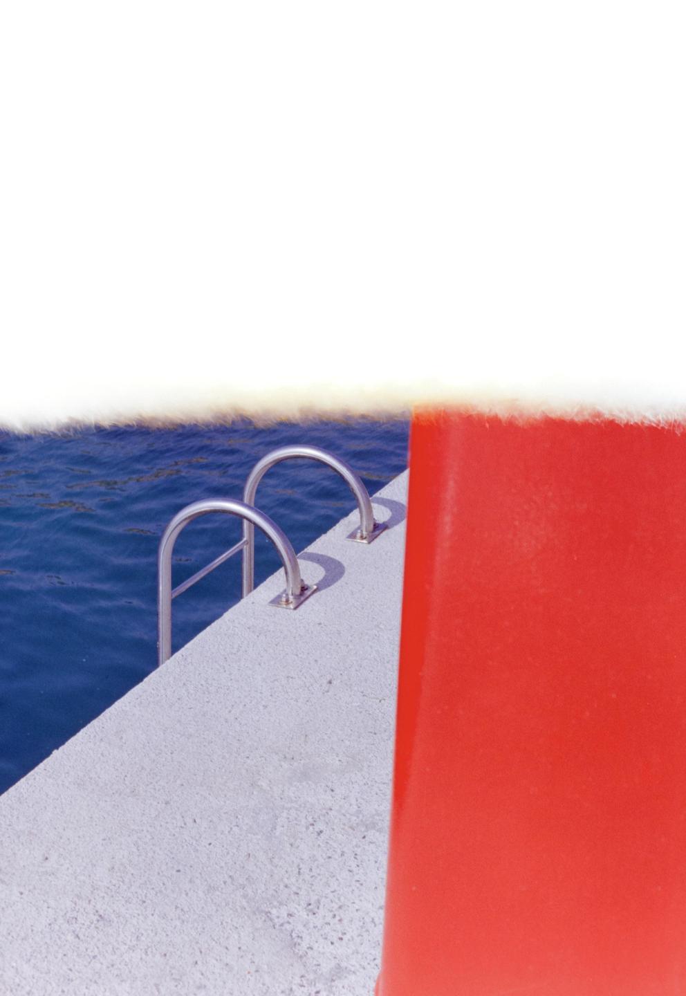 A metal ladder extends into blue water from a concrete edge next to a red surface.