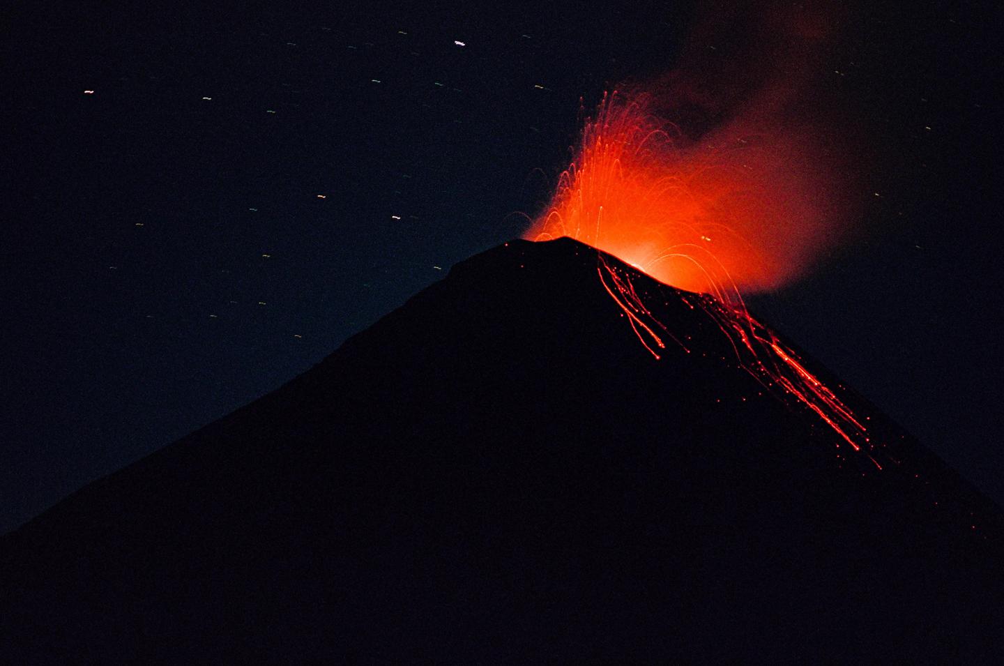 A volcano erupts at night, spewing bright orange lava and smoke into the dark sky.