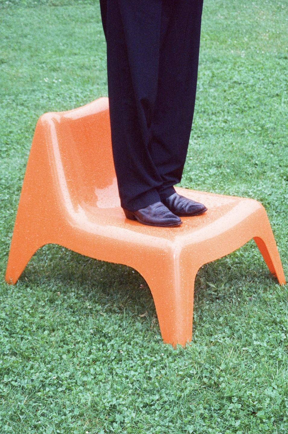 A person stands on an orange plastic chair in a grassy area.