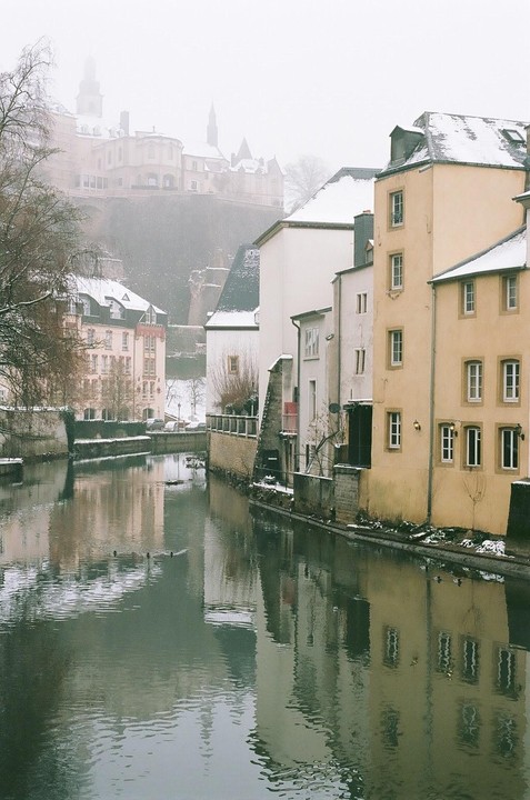 A river flows between buildings with snow-covered roofs and a distant castle on a hazy winter day in Luxembourg.