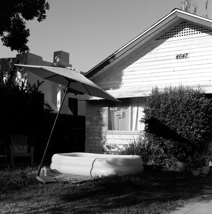 An inflatable pool sits in a grassy yard next to a house with an open umbrella providing shade.