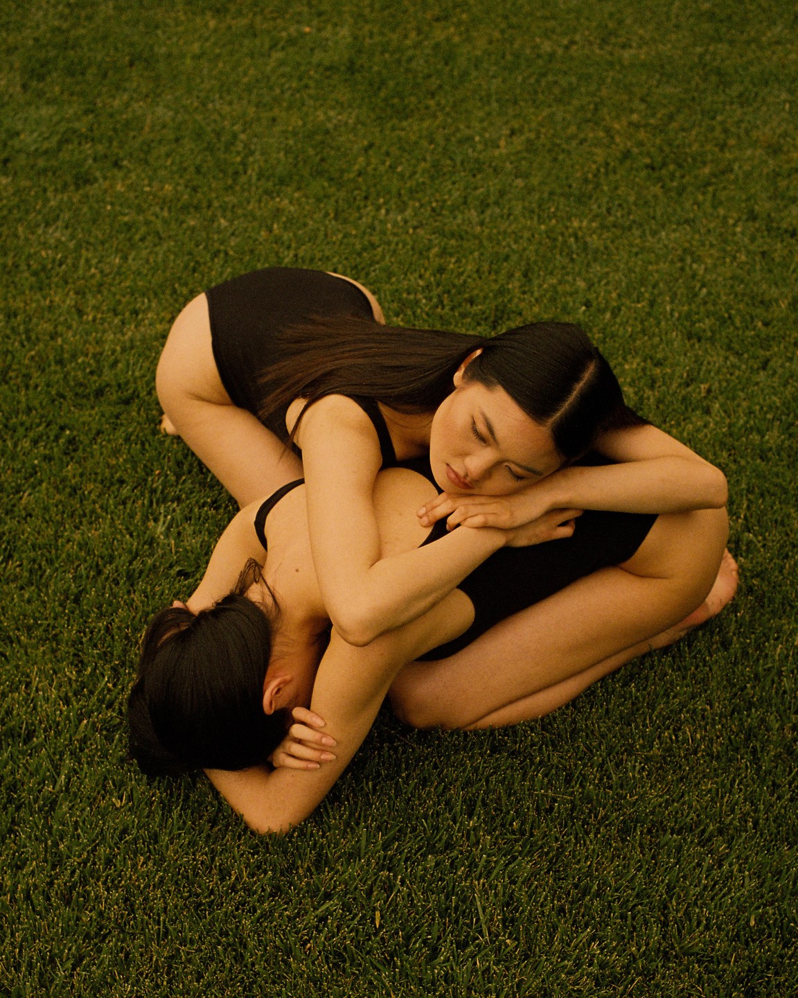Two women in black leotards are intertwined on a patch of green grass.