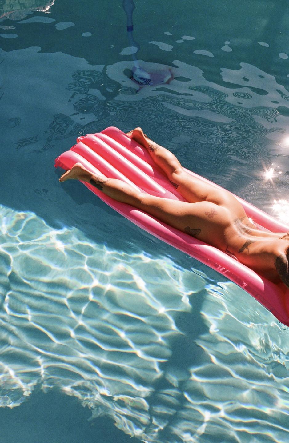 A person with tattoos floats on a pink inflatable raft in a swimming pool.