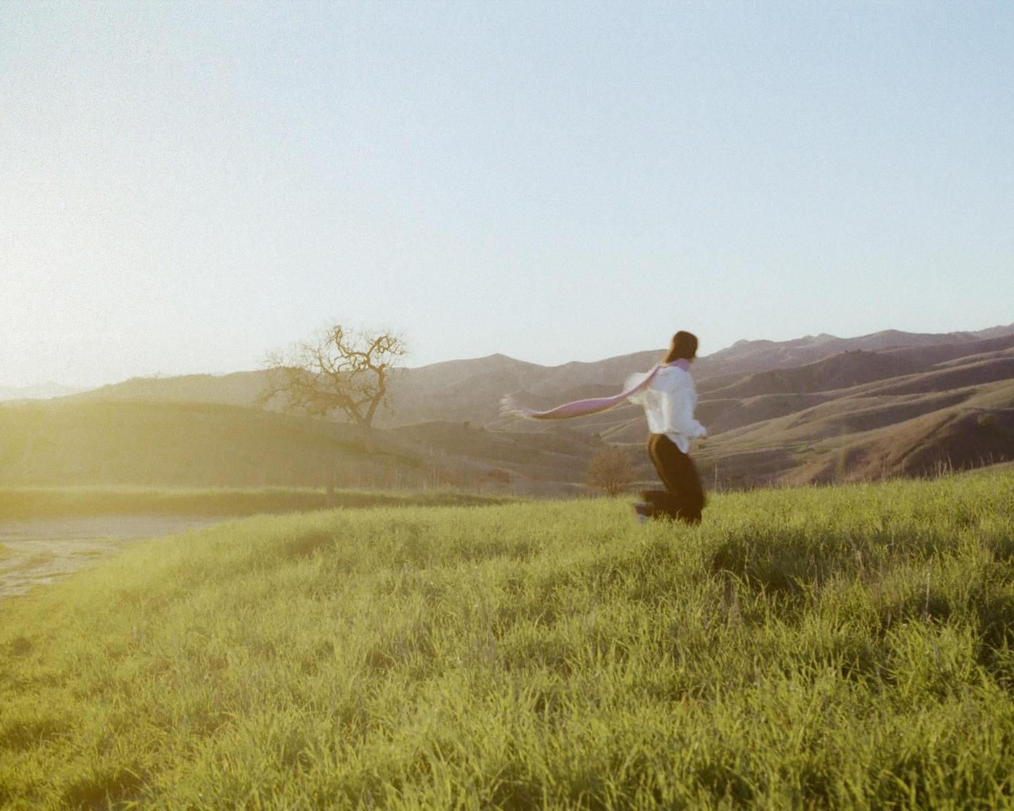 A woman jumps in a grassy field with rolling hills and a tree in the background.