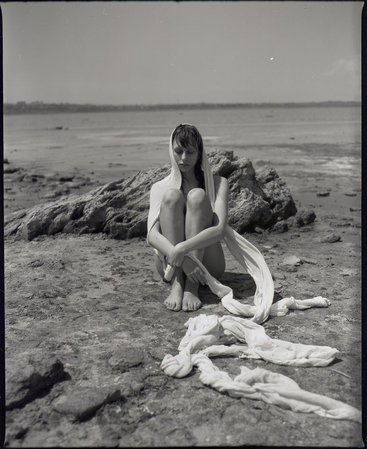 A woman sits on a rocky shore with a white cloth draped over her head and shoulders.