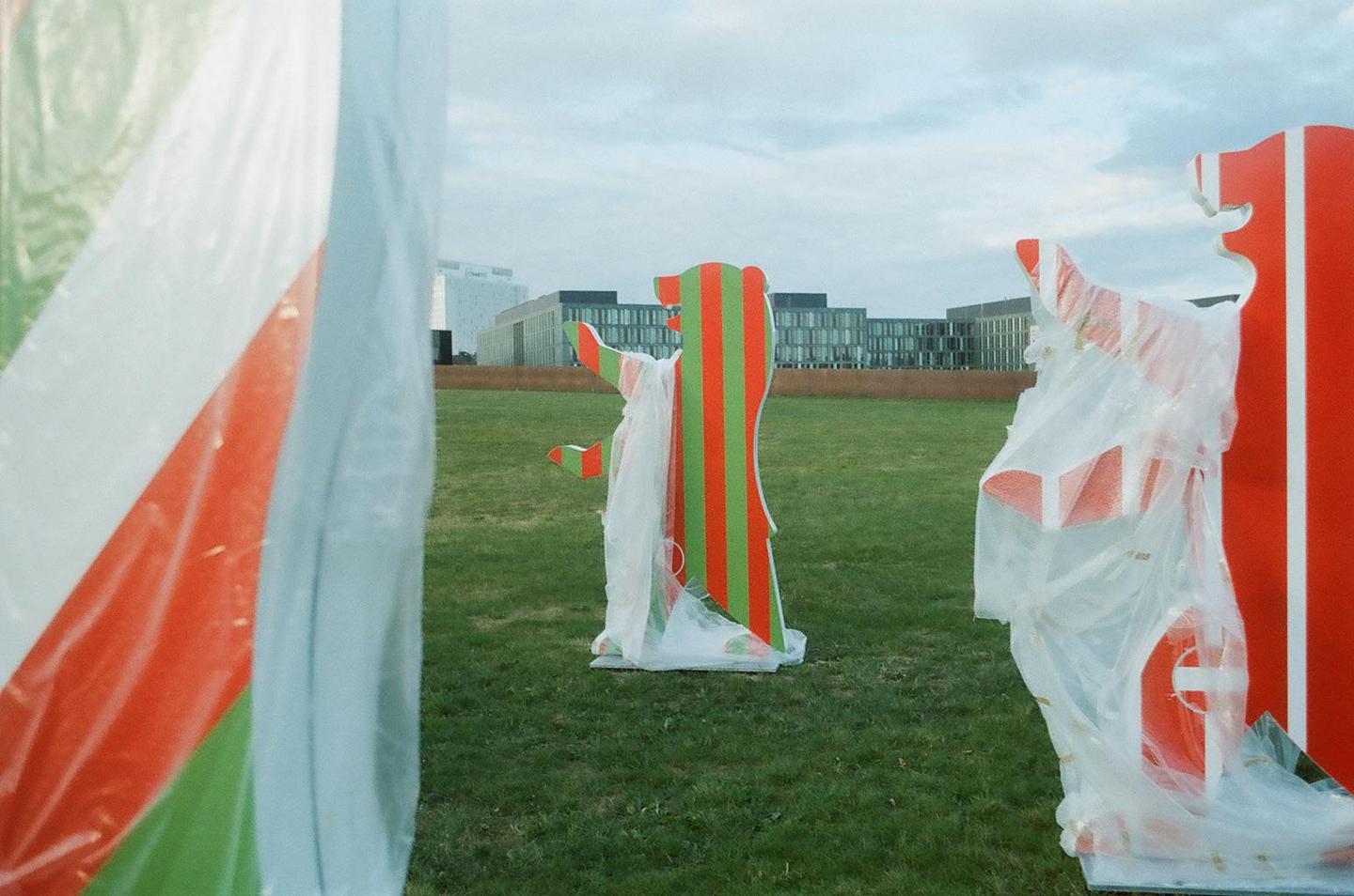 A red and green striped bear statue stands in a grassy field with buildings in the background.