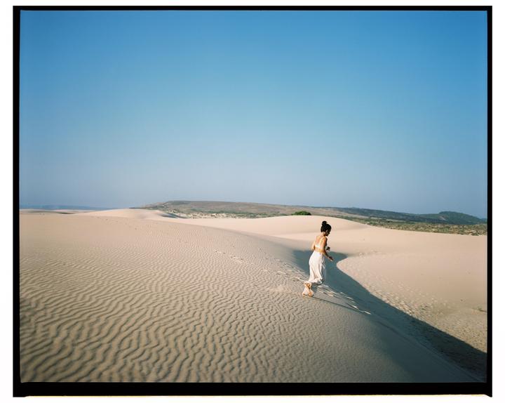 A woman walks barefoot on a sand dune under a clear blue sky.