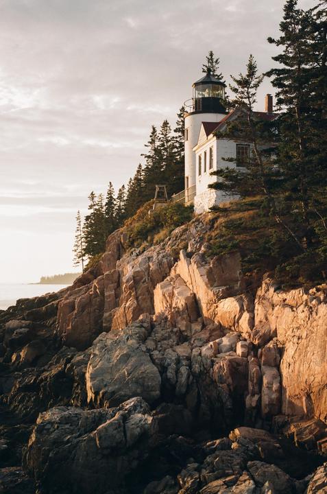 A white lighthouse and house stand on a rocky cliff overlooking the ocean at golden hour.