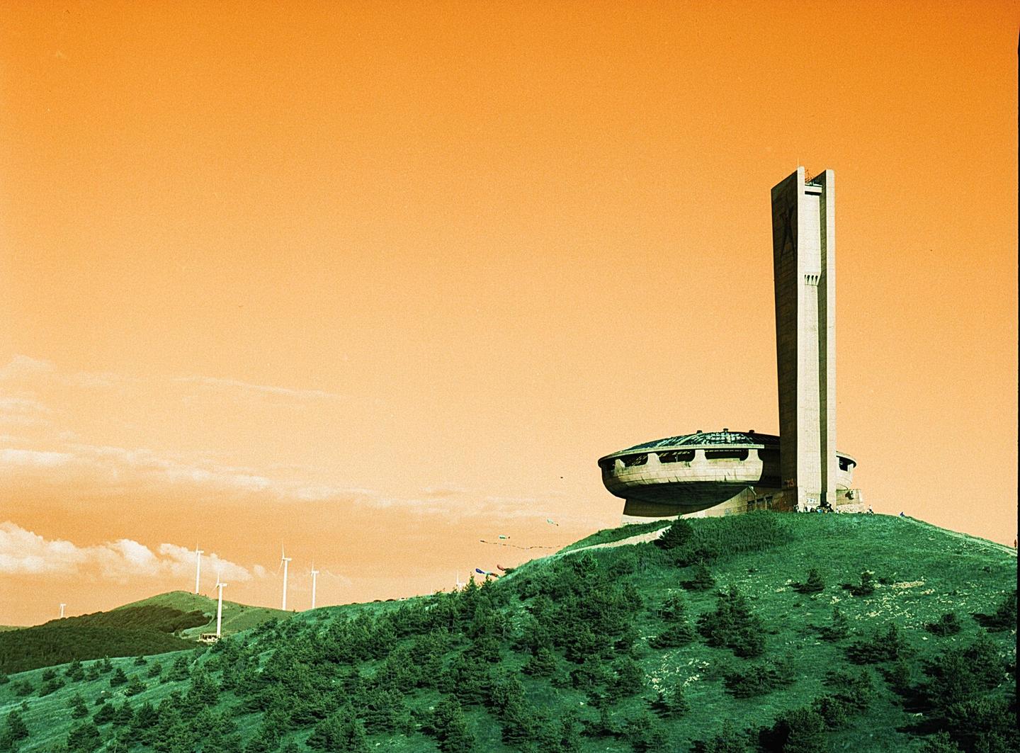 A brutalist monument with a tall tower and a circular base sits atop a green hill under an orange sky.