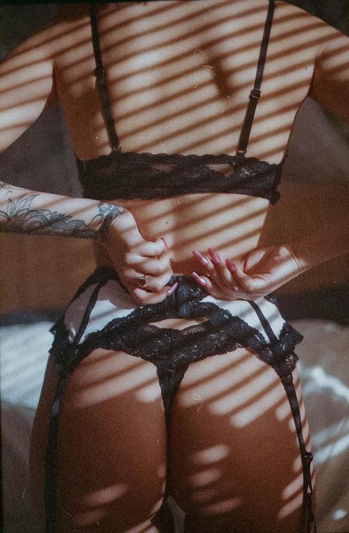 A woman in lingerie is seen from behind, fastening her garter belt, with shadows from blinds cast across her body.