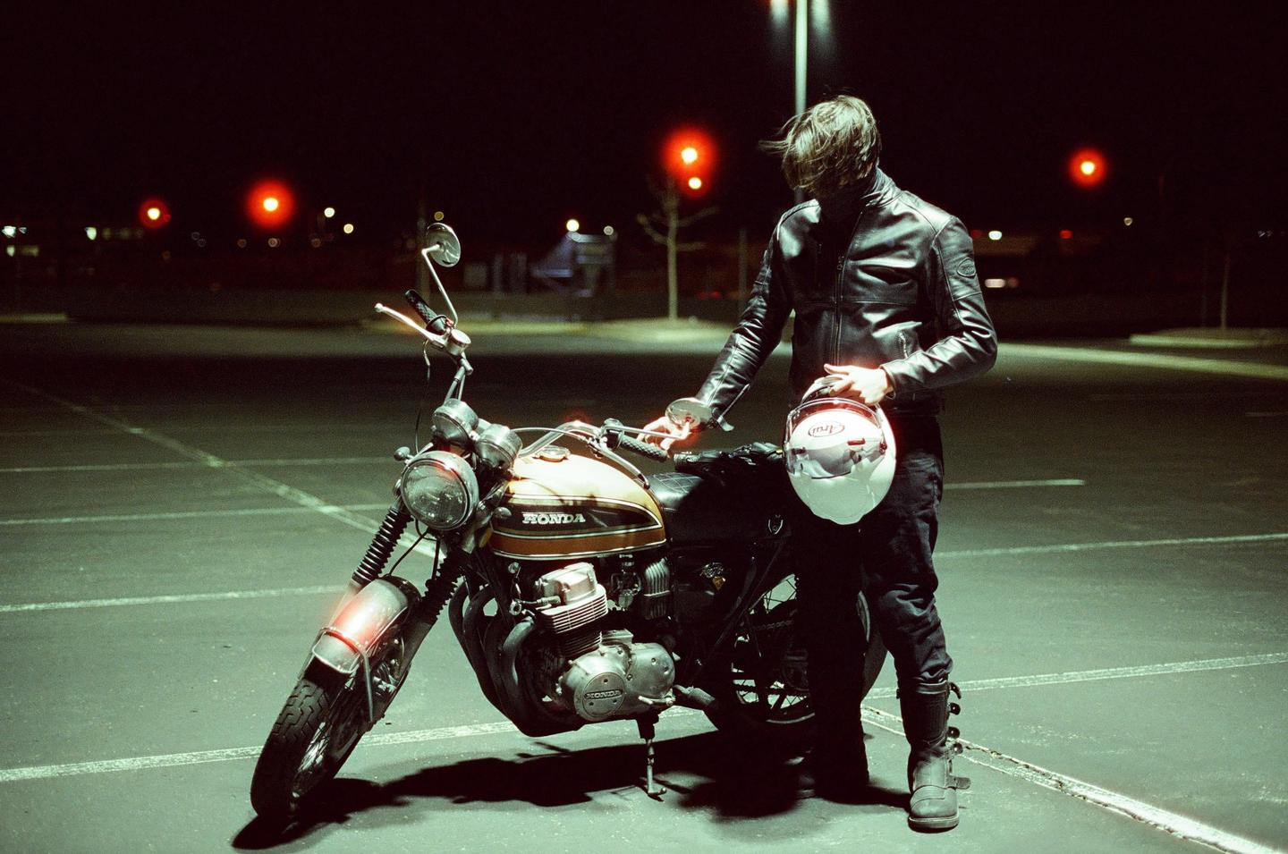 A man in a leather jacket stands next to a motorcycle at night, holding a helmet.